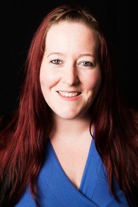 Heather, a white femme with pale skin and long red hair is wearing a blue top on a back blackground