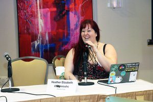 Heather sitting at the table with her laptop and a coffee cup in front of them. She is speaking into a microphone